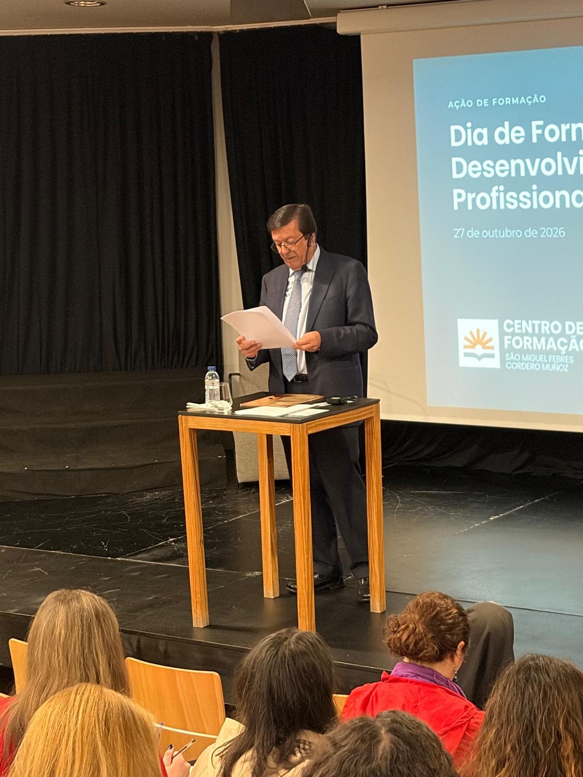 Orador a ler um texto no palco, diante do ecrã do Dia de Formação e Desenvolvimento Profissional