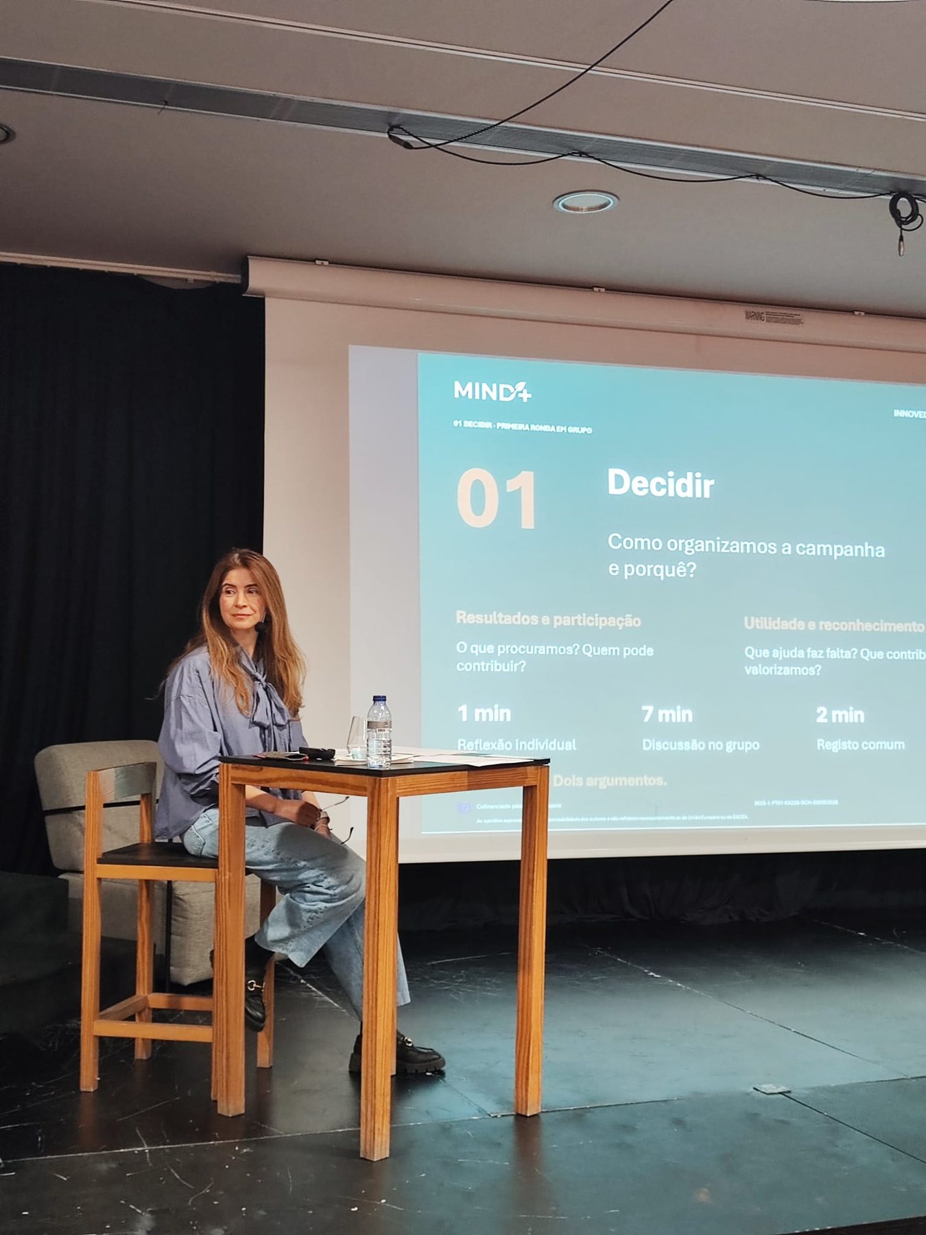 Formadora sentada no palco, diante de um diapositivo com a proposta de trabalho em grupo