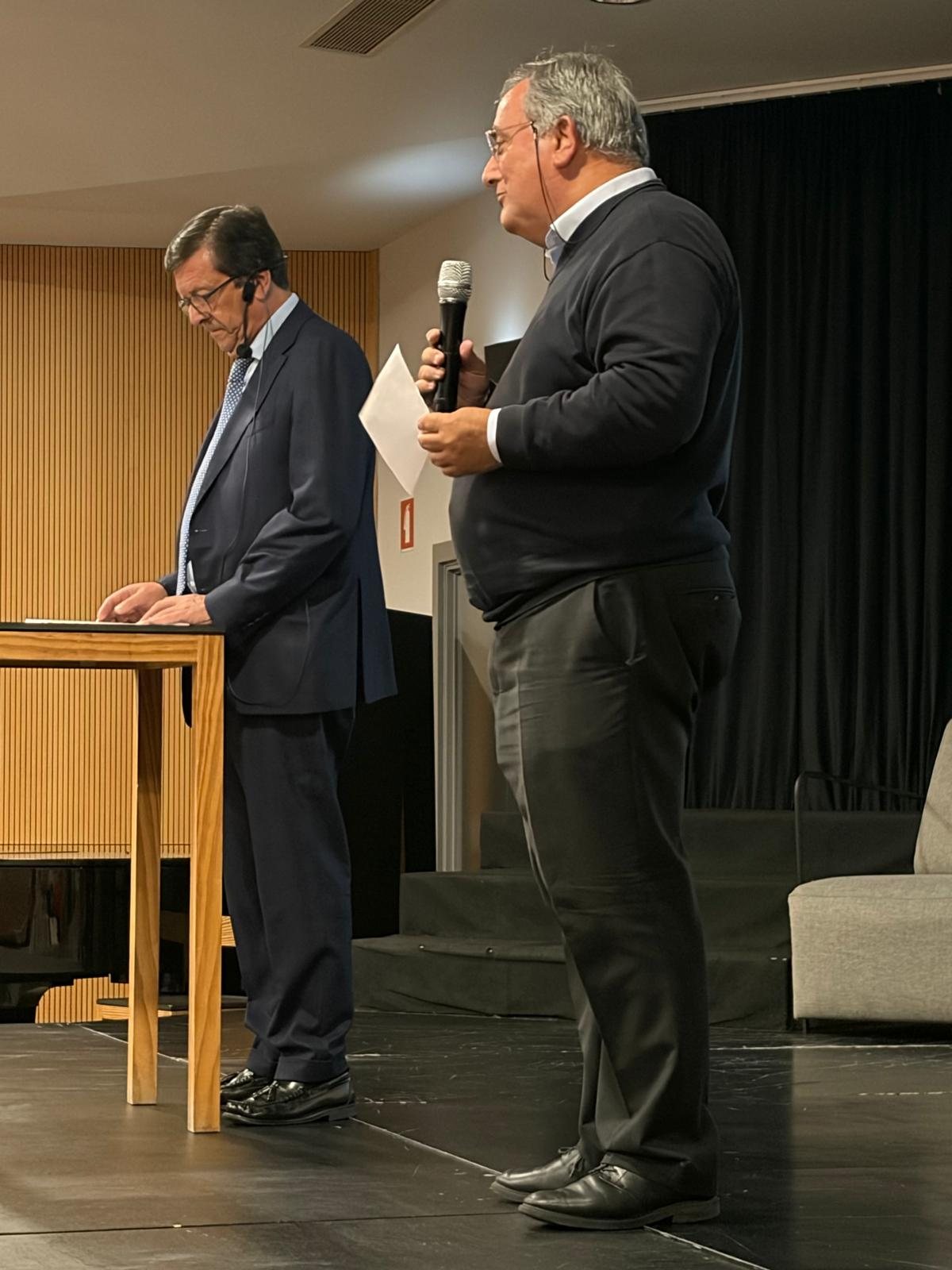 Dois oradores no palco, um a ler e outro de microfone na mão