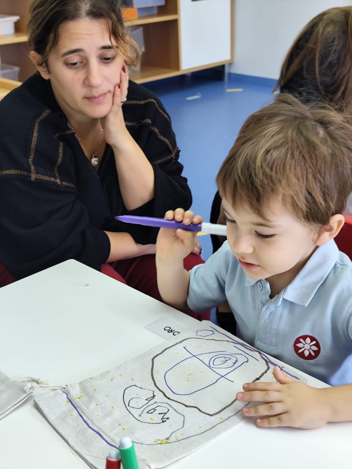 Criança a desenhar no seu saquinho de pano, com a mãe ao lado